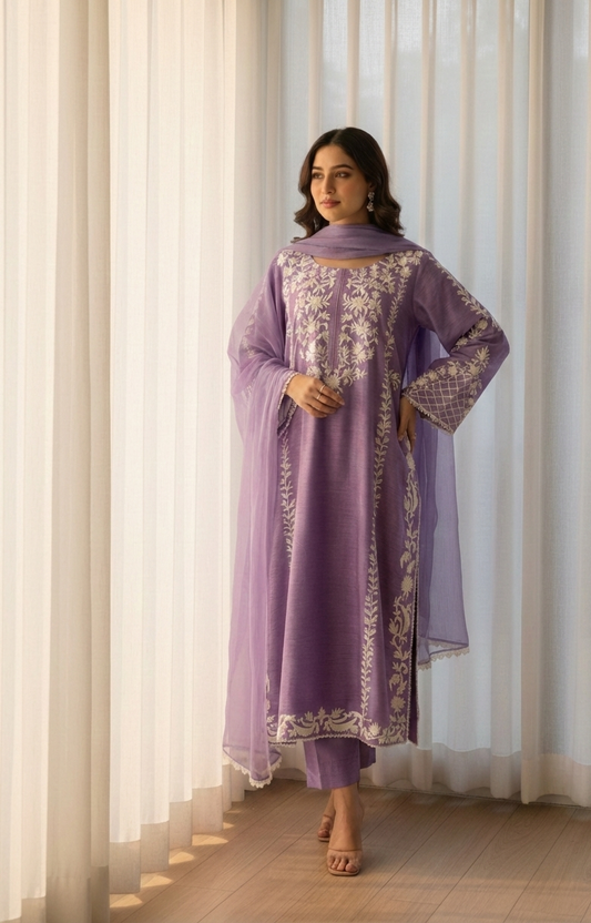 Woman wearing a purple traditional outfit with white embroidery standing in front of sheer curtains.